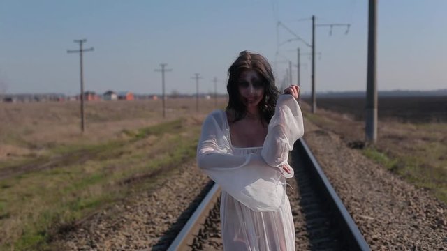 Portrait amazing slim unusual girl in white translucent dress and with makeup on her face in form of torn bloody wound makes unusual dance moves on railway tracks in sunny day