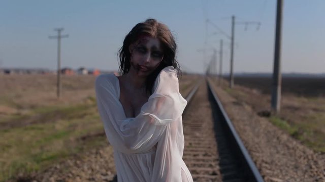 Portrait amazing slim unusual girl in white translucent dress and with makeup on her face in form of torn bloody wound makes unusual dance moves on railway tracks in sunny day