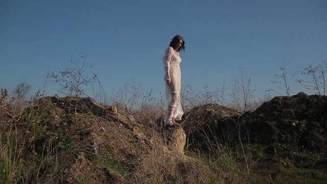 Camera motion, amazing slim unusual girl in white translucent dress and with wax makeup on face in form of bloody wound walks in field and makes beautiful unusual dance moves under boundless blue sky