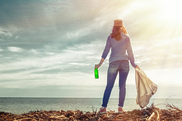 The woman volunteer stands on the shore of the sea. She holds a garbage bag in one hand and a dirty...