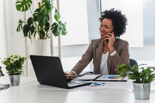 Happy Smiling African-american Business Woman Working On Laptop At Office, Using Smart Phone. Businesswoman Sitting At Her Working Place