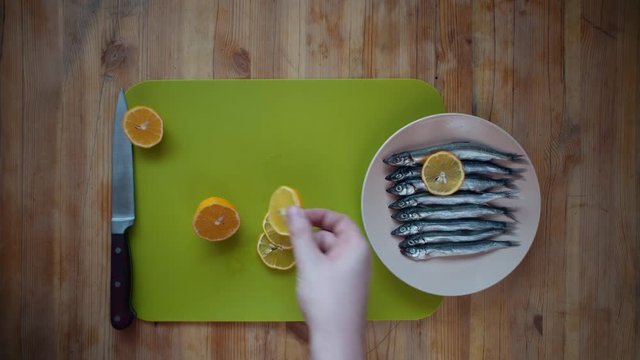 Top view of the process of making a small silver fish, male hands cover capelin with fresh lemon slices, lying in a beige plate on a wooden table.