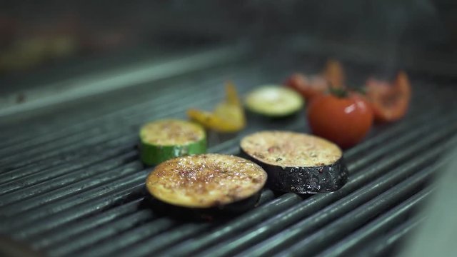 Close up of chef hand puts vegetables tomato, eggplant, yellow bell pepper on the grill using metal tongs. Cook preparing vegetarian food. Healthy lifestyle