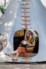A beautiful girl in an ethnic Indian costume and in the stream sits on the threshold of the tipi....
