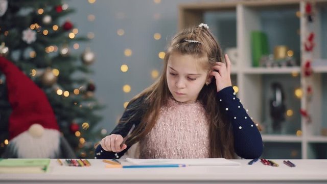 Girl learning lessons, cute girl making her homework, schoolgirl studing drawing at home at the table, children's education, back to school. Christmas tree on the background. Winter holidays, positive