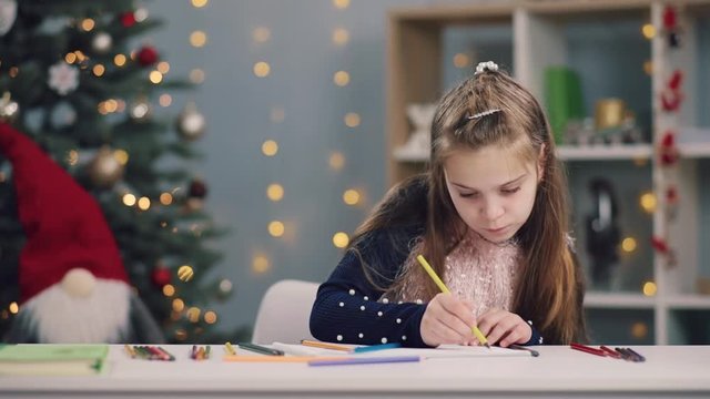 Portrait of cute little girl drawing with a yellow pencil then changing it on a blue one on Christmas tree lights background. Creativeness, talent, art class. Art therapy, holidays. Child�s portrait