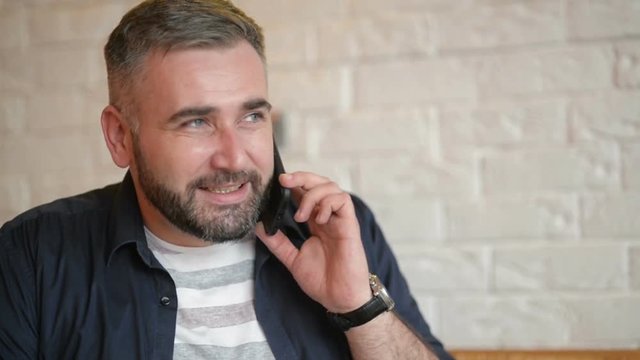 Handsome Businessman Talking on the Phone at the Cafe. Young Bearded Guy Holding His Smartphone Near His Ear. Attractive Caucasian Man in White Shirt Calling Somebody On His Cellphone.