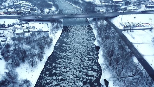 Forward drone shot, panoramic view, of the river Neris, Vilnius, Lithuania, flowing with a frozen ice surface, surrounded by snow and trees, near the city and a bridge with cars, on a winter day