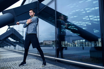 Active man doing his stretching workout next to modern architecture with glass wall. Urban area...