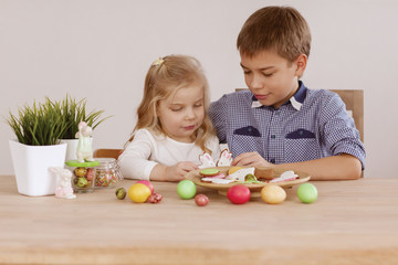 Obraz premium A girl with an older brother are sitting at the holiday table and laying out cookies and Easter eggs