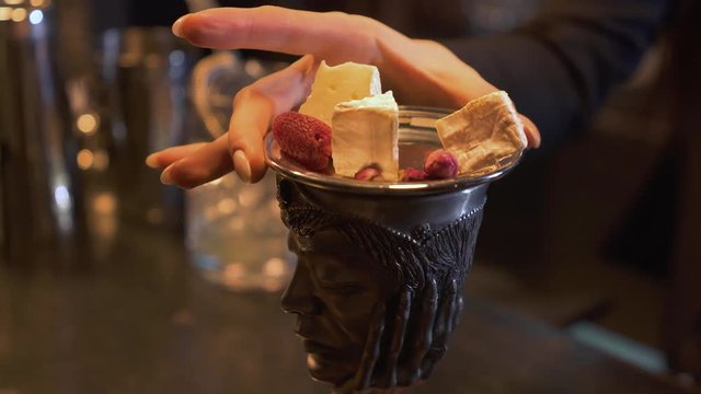 Metal cup with beverage on the bar. Woman put on top of bowl metal sauser with pieces of cheese, dry roses and raspberry