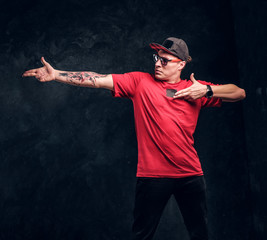 Stylish man with a tattoo on his hand dressed in a hip-hop style, posing for a camera. Studio photo against a dark textured wall