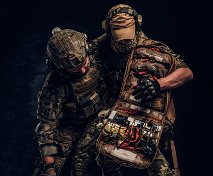 Combat Conflict, Special Mission, Retreat. Military Medic Rescues His Wounded Teammate Carrying Him Off The Battlefield. Studio Photo Against A Dark Wall