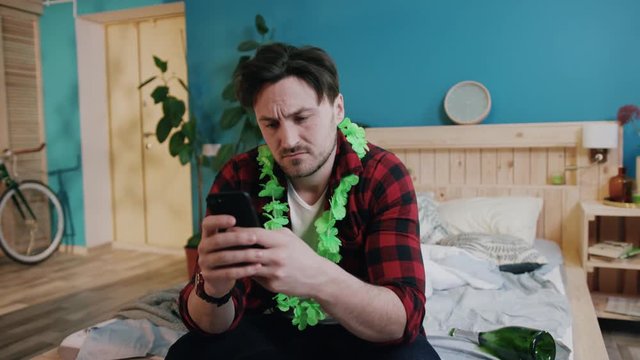 Serious Caucasian man in plaid shirt holding black smartphone in hand. Guy hard to concentrate and execute fine motor skills. Indoors. Unpleasant experience of heavy drinking.