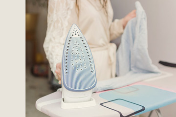 beautiful young woman ironing clothes on an ironing board smiles looking at the camera, being at home against the window in which the sun shines