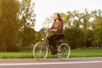 Fototapeta na wymiar Young attractive woman riding through the park after work. Beautiful lady cycling during sunset. Bike as a trendy transport. Healthy outdoors activity on a warm summer day. Bicycle trend in the city.