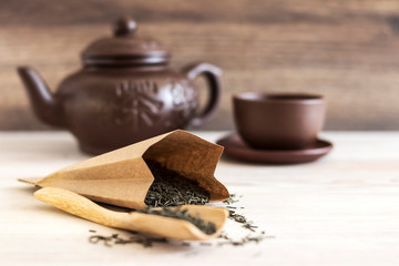 Wooden spoon with green tea and tea bag against a brown clay teapot and cup