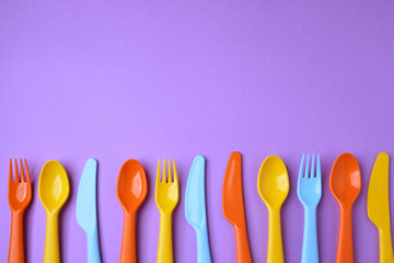 Many colorful plasic forks, spoons and knives on violet background with copy space, top view.