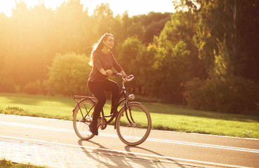 Young attractive woman riding through the park after work. Beautiful lady cycling during sunset. Bike as a trendy transport. Healthy outdoors activity on a warm summer day. Bicycle trend in the city.