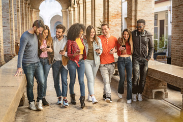 Obraz premium young adults converse and joke together using their smartphones walking under the arcades of a European city. Social media concept.