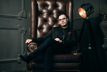 Obraz na płótnie Canvas Young handsome man posing for a fashion shoot in a studio. Fashion as a lifestyle. Man wearing a beard. Model sitting on a sofa in tuxedo. Successful fashionable businessman. Business look advertising