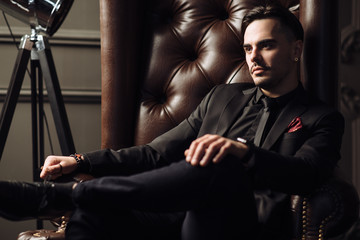 Young handsome man posing for a fashion shoot in a studio. Fashion as a lifestyle. Man wearing a beard. Model sitting on a sofa in tuxedo. Successful fashionable businessman. Business look advertising