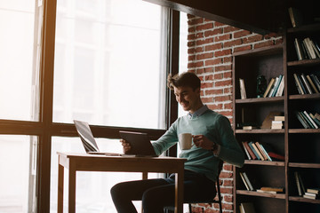 Young handsome man sitting in office with cup of coffee and working on project connected with modern cyber technologies. Businessman with notebook trying to keep deadline in digital marketing sphere.