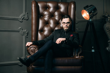 Fototapeta na wymiar Young handsome man posing for a fashion shoot in a studio. Fashion as a lifestyle. Man wearing a beard. Model sitting on a sofa in tuxedo. Successful fashionable businessman. Business look advertising