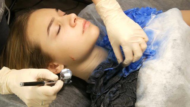 Fashion studio, the model is preparing for the show. Teen girl lies on a special couch, the designer prepares her image using a special airbrush spray with paint