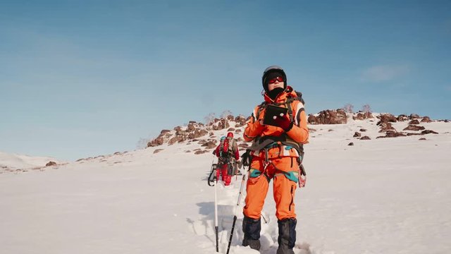 girl climber in an orange suit stands with an electronic tablet with a map in his hands and checks the route. in the background are a mountain and three climbers.