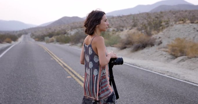 Stylish female photographer walking on desert highway with camera - slow motion