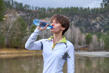 Fitness woman is drinking water from a bottle on a landscape background. Outdoor sports portrait