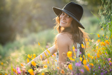 Beautiful young latino woman in a hat smiling while sitting back to camera in poppy fields at sunset