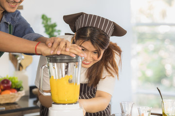 Close up portrait of happy young Asian couple helping prepare orange smoothie with electric juice blender, in modern kitchen space with large glass window and blur background