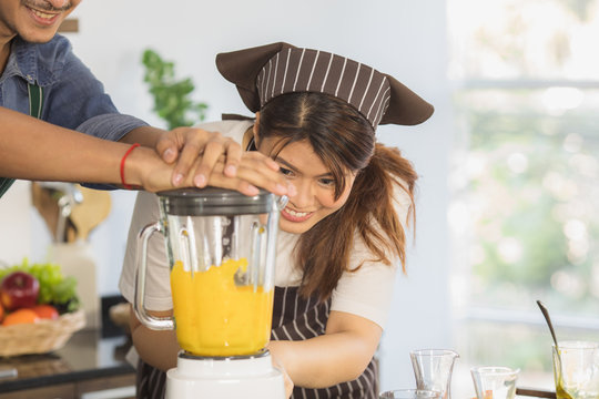 Close up portrait of happy young Asian couple helping prepare orange smoothie with electric juice blender, in modern kitchen space with large glass window and blur background - Powered by Adobe