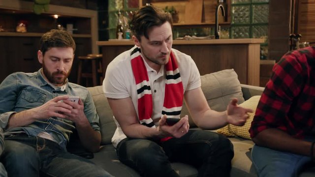 Attractive mixed race men hanging out at home, drinking beer and eating snacks while using their smartphones all at the same time, zoom in view of bearded man showing his friends his smartphone screen