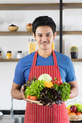 Close up shot of happy young Asian handsome bearded man wearing blue t shirt and striped apron hold a plate full of vegetables, presenting healthy meals images standing with modern kitchen background