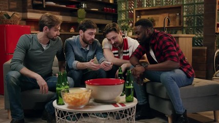Four young male friends using smartphone, man in the center shows the screen of the phone, the rest of the company dismiss him and leave the room, bearded attractive young man sits alone surprised - Powered by Adobe