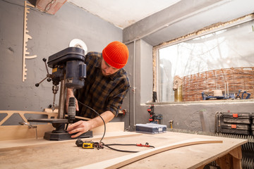 A man with work clothes and a carpenter's cap is carving a wooden board on an modern large drilling machine in a light workshop