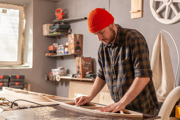 A young man in a orange hat  carpenter builder in work clothes processing a wooden board with a milling machine in the workshop, around a lot of equipment, wooden boards