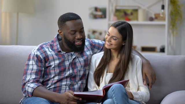Cheerful mixed-race couple viewing family photo album at home, pleasure memories
