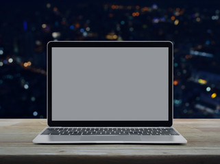 Modern laptop computer with blank screen on wooden table over blur colorful night light office city tower and skyscraper