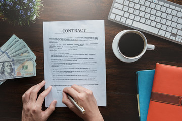 contract and agreement concept, man holding pen to sign contract application paper with coffee cup keyboard dollar bank and notebook on wood desk in office bank.