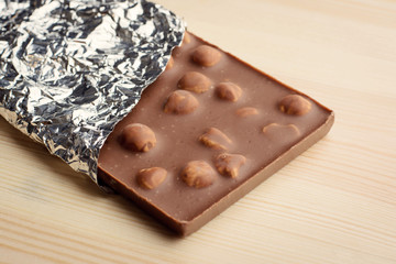 Bar of chocolate with hazelnuts and covered with foil lays on light wooden background.
