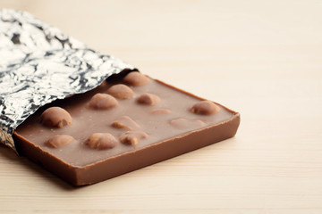 Bar of chocolate with hazelnuts and covered with foil lays on light wooden background.