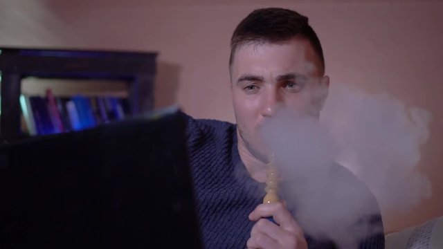 The young guy smokes a hookah, exhales smoke and works at a laptop. A young man in the smoke. A young guy on a pillow against a background of a bookcase and works at a laptop.