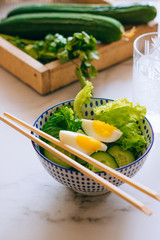 Healthy bowl, cucumber salad with eggs, coriander in blue plate and cucumber in wooden box on marble background