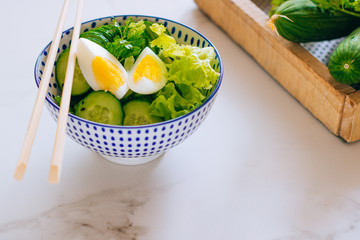 Healthy bowl, cucumber salad with eggs, coriander in blue plate and cucumber in wooden box on marble background