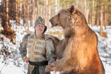 A man in traditional national costume of a Russian prince stands by a brown bear in a winter...
