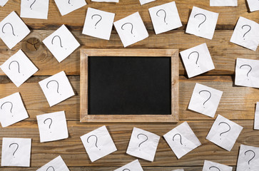 Question marks on crumpled note paper around a small blank blackboard on a wooden background